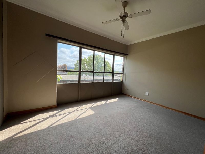 A spacious, empty bedroom featuring neutral carpeting, a ceiling fan, and a large window providing natural light and an exterior view.