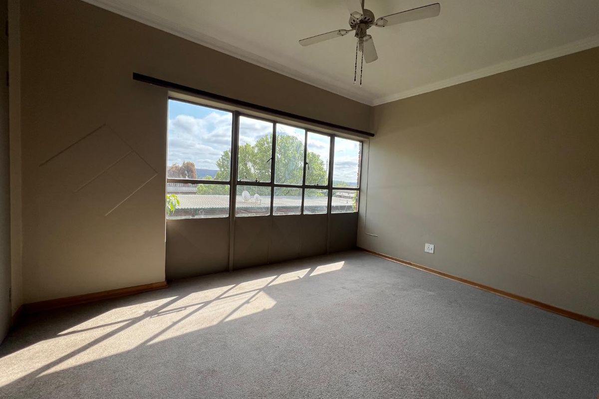 A spacious, empty bedroom featuring neutral carpeting, a ceiling fan, and a large window providing natural light and an exterior view.