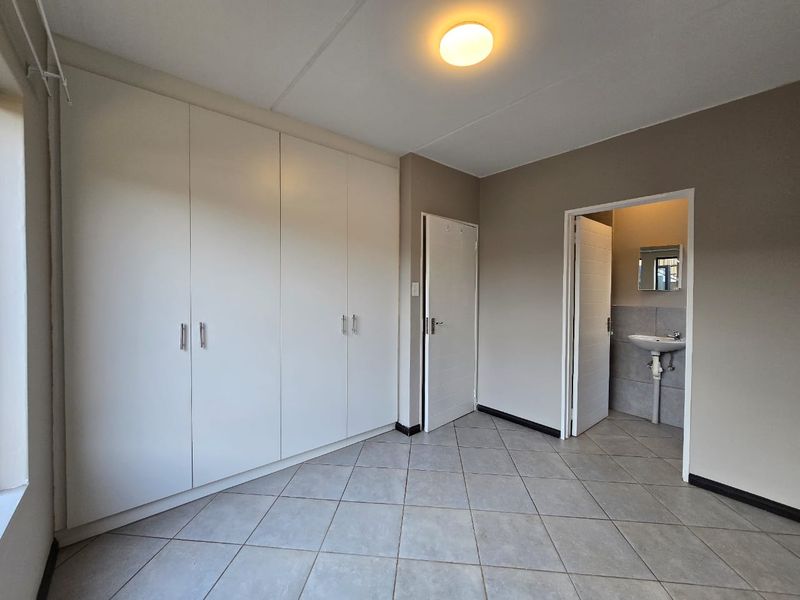 A clean, empty bedroom featuring built-in cupboards, tiled flooring, and an attached en-suite bathroom with a pedestal basin.