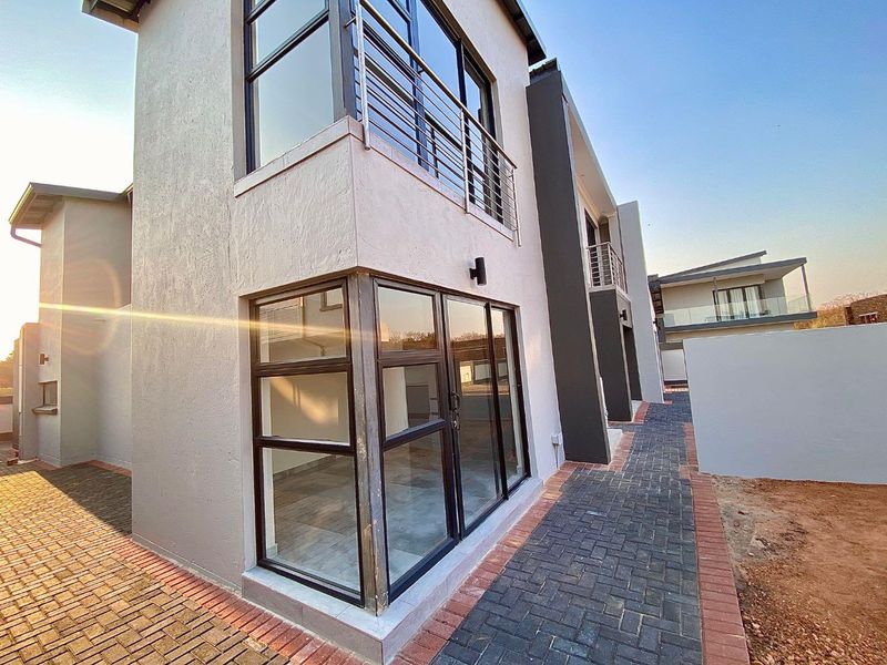 Modern double-story residential exterior featuring paved walkways, large aluminium-framed windows, and an upper-level balcony with metal railings.