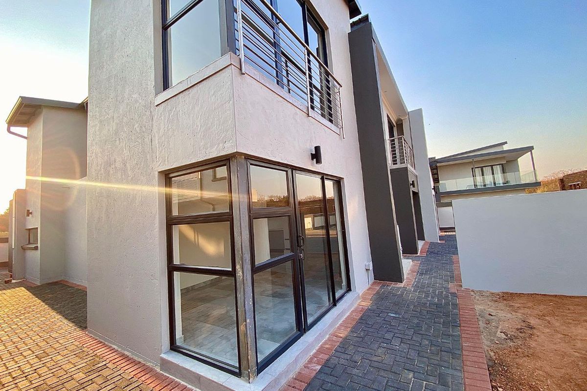 Modern double-story residential exterior featuring paved walkways, large aluminium-framed windows, and an upper-level balcony with metal railings.