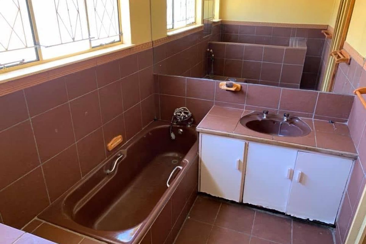A dated bathroom featuring brown tiled walls and flooring, a built-in bathtub, and a vanity unit with a large wall-mounted mirror.