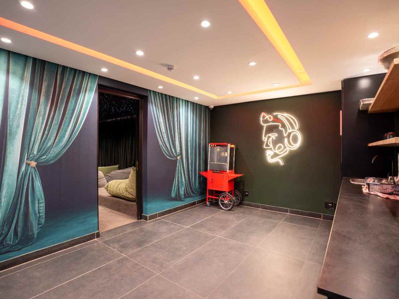 A modern cinema room featuring recessed ceiling lighting, cove lighting, tiled flooring, and a convenient kitchenette area with built-in shelving and a sink.