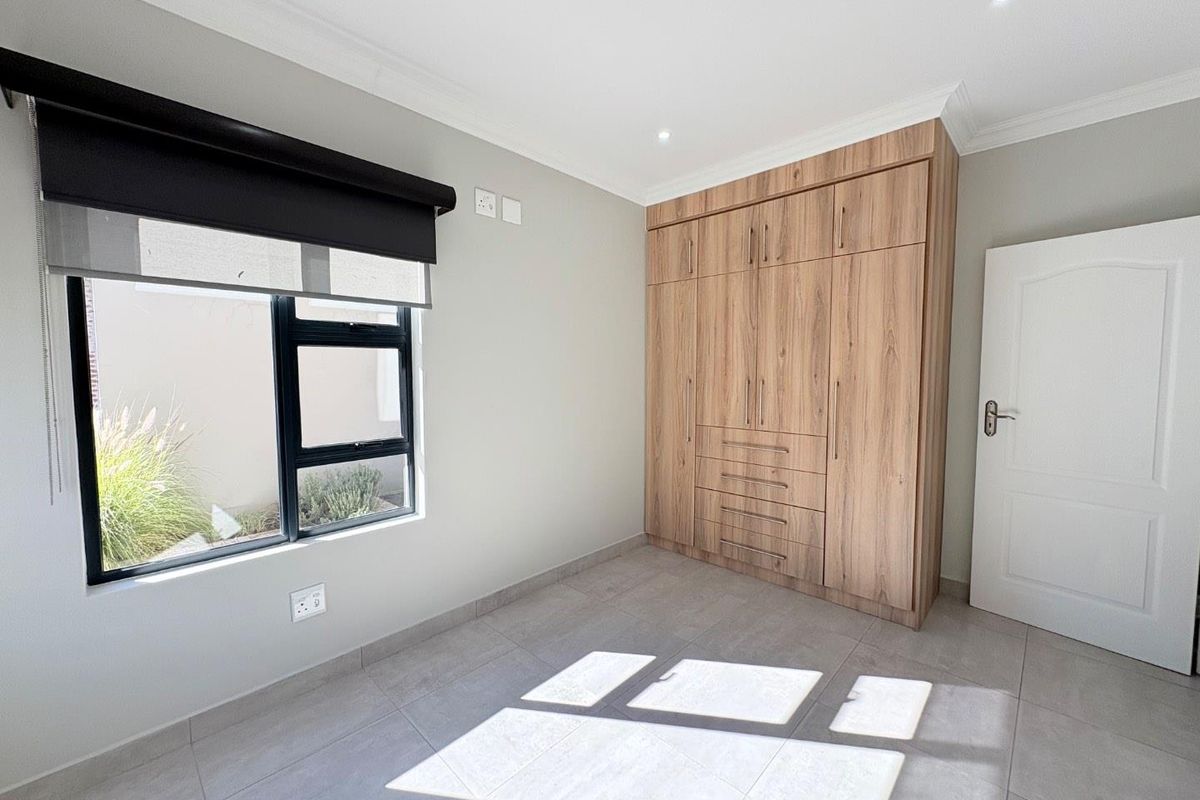 A bright, modern bedroom featuring neutral floor tiles, built-in wooden cabinetry, and a large window with a roller blind.