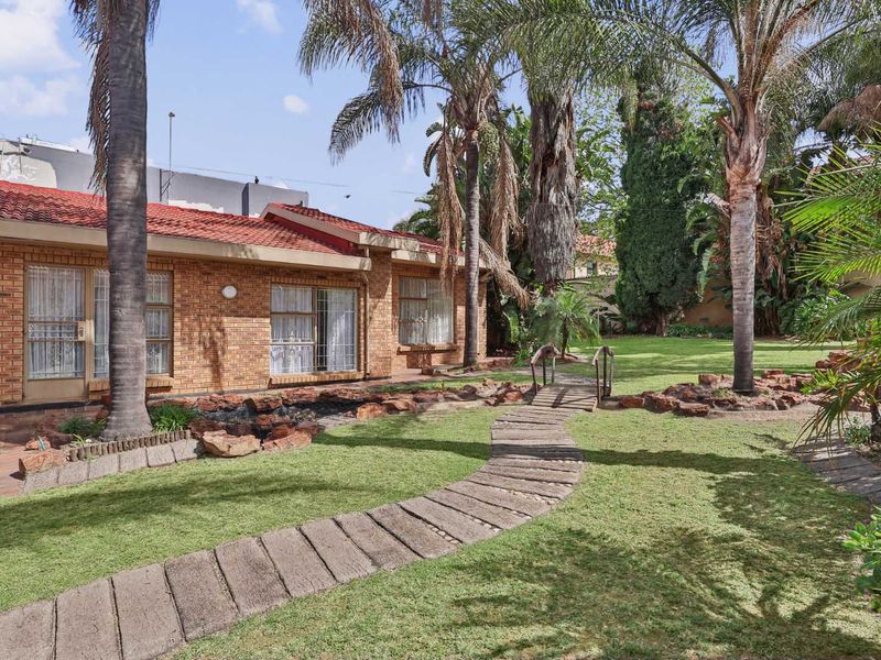 A well-maintained rear garden featuring a brick facade house, a paved walkway with a small wooden footbridge, and established palm trees.