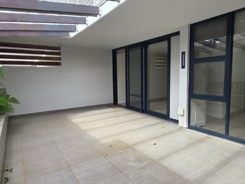 A spacious covered patio featuring tiled flooring, a timber pergola, and a built-in planter box, with large sliding glass doors leading into the apartment.