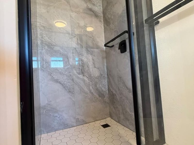 Modern walk-in shower featuring marble-look wall tiles, hexagonal floor tiles, and contemporary black fixtures including a rain shower head and glass screen frame.