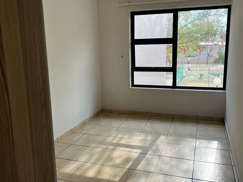 A bright, empty bedroom featuring neutral wall tones, tiled flooring, and a large aluminium-framed window providing natural light.