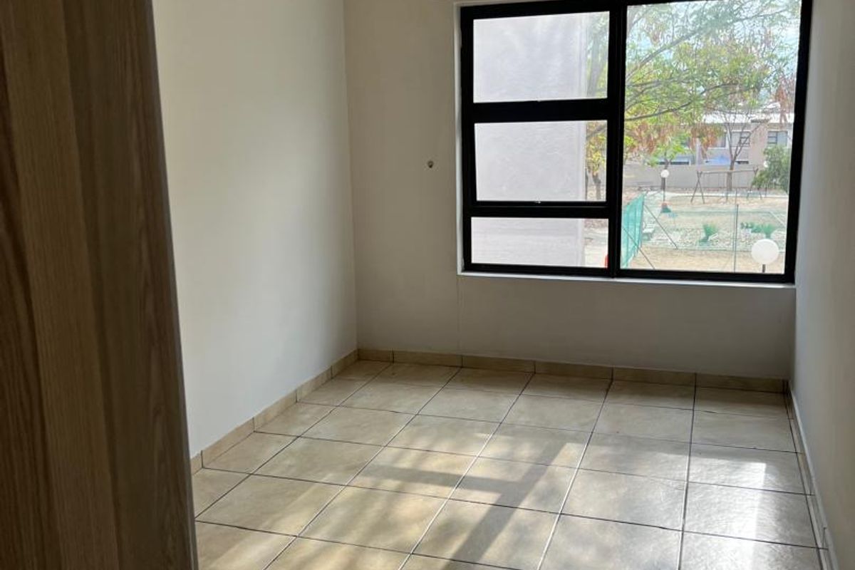 A bright, empty bedroom featuring neutral wall tones, tiled flooring, and a large aluminium-framed window providing natural light.