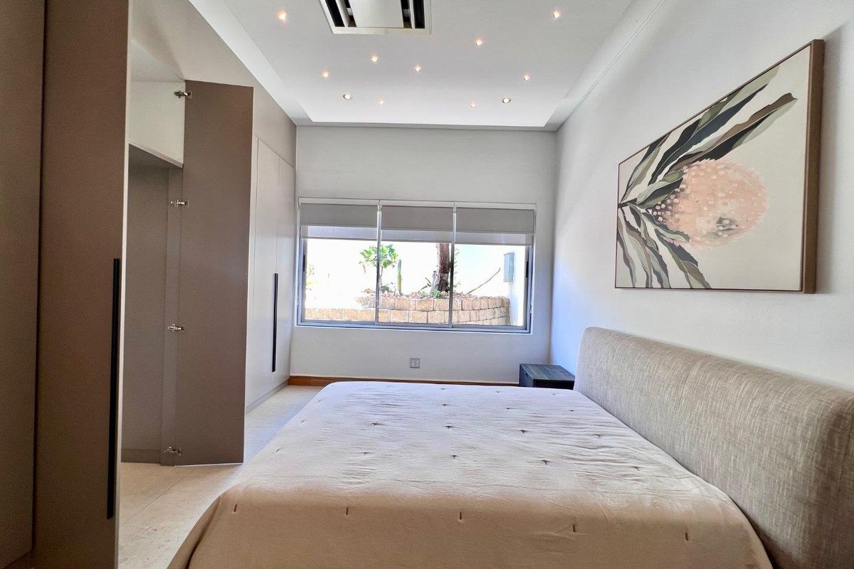 A modern, well-lit bedroom featuring built-in floor-to-ceiling cabinetry, recessed ceiling lighting, and large windows overlooking a landscaped garden area.