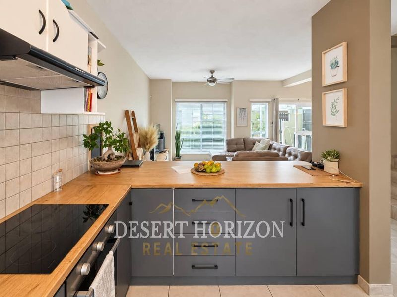 Open-plan kitchen featuring modern grey cabinetry, wooden countertops, and integrated appliances, flowing seamlessly into a bright living area with large windows.