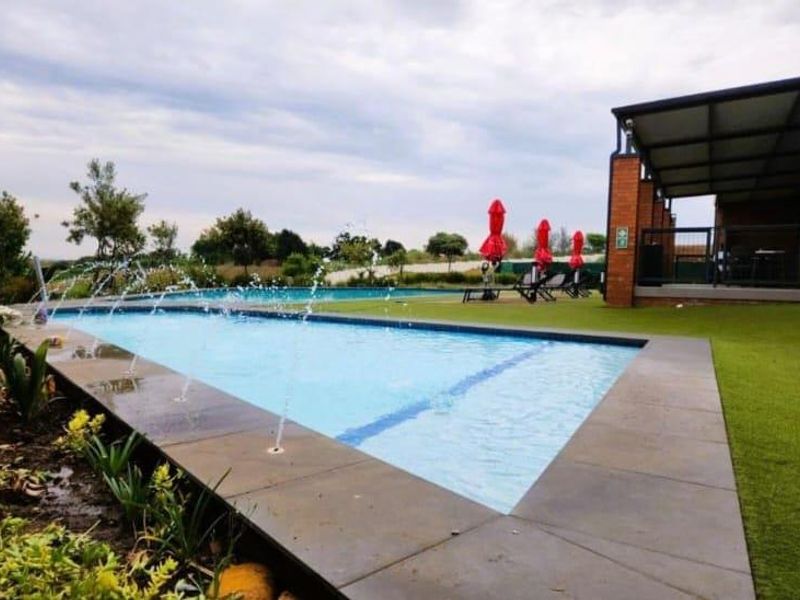 A well-maintained outdoor pool area featuring a rectangular swimming pool with water fountains, adjacent to a covered patio and landscaped artificial turf.