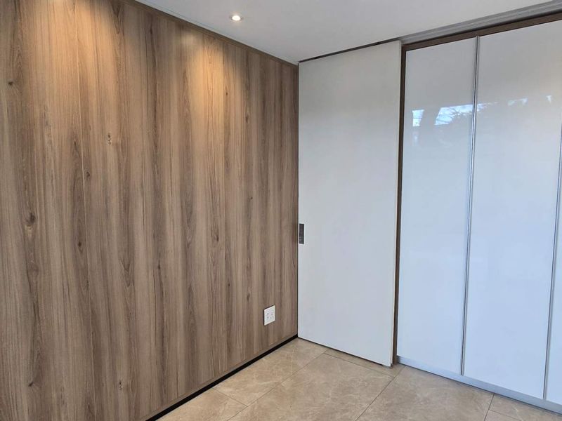 A modern, minimalist bedroom featuring high-quality wood-look wall paneling, sleek built-in cupboards, and neutral tiled flooring with recessed lighting.