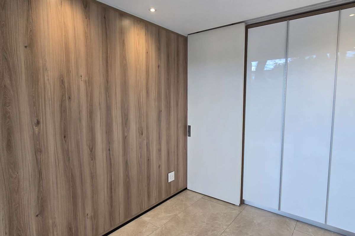 A modern, minimalist bedroom featuring high-quality wood-look wall paneling, sleek built-in cupboards, and neutral tiled flooring with recessed lighting.