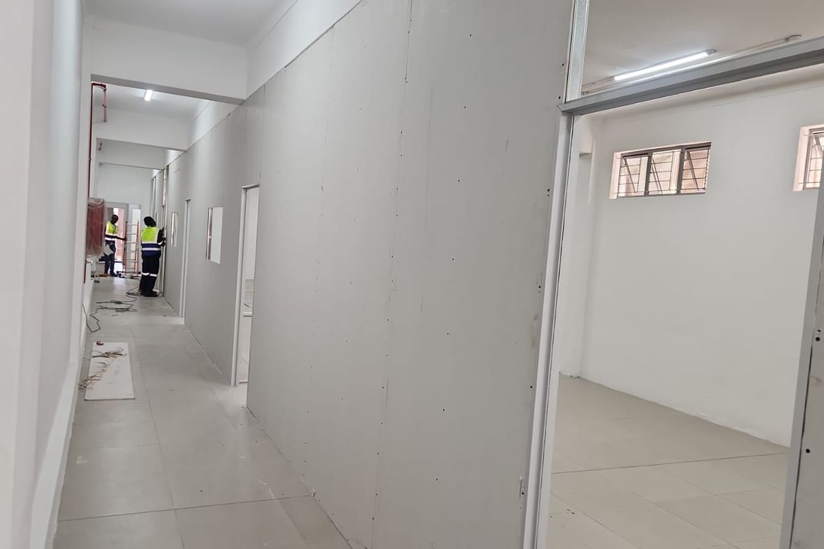 A hallway in an office space currently under construction, featuring unfinished drywall partitions, tiled flooring, and overhead lighting.