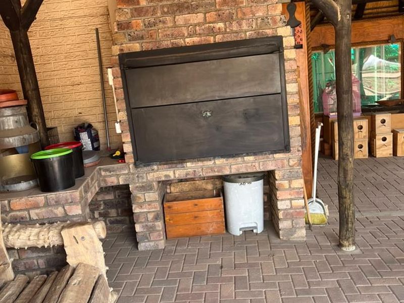 Covered outdoor patio area featuring a built-in brick braai, rustic timber support poles, and durable brick paved flooring under a traditional thatch roof.