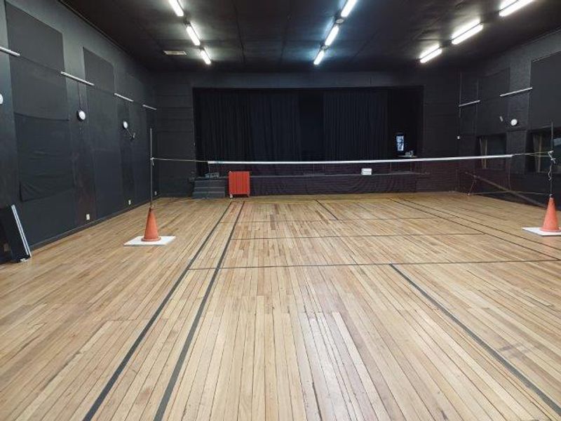 A large multi-purpose sports facility featuring polished hardwood flooring, a raised stage area, and acoustic wall paneling, suitable for various recreational or office activities.