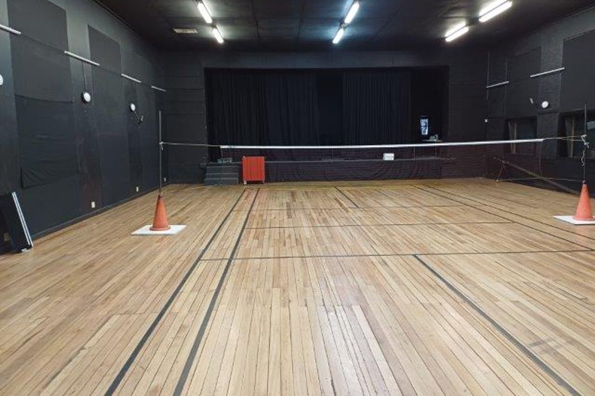 A large multi-purpose sports facility featuring polished hardwood flooring, a raised stage area, and acoustic wall paneling, suitable for various recreational or office activities.