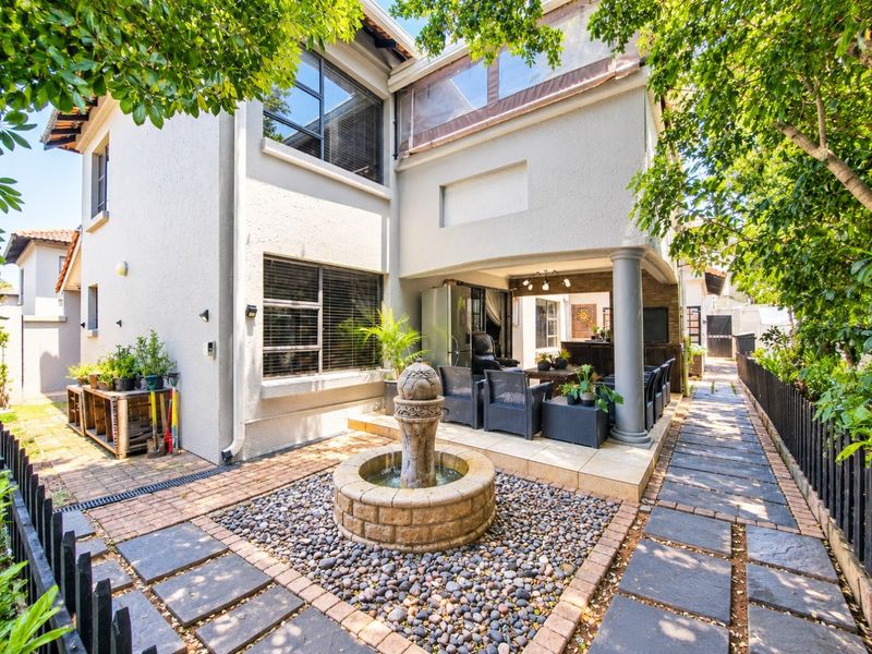 A well-maintained rear exterior featuring a covered patio, a decorative stone fountain, and paved pathways set within a landscaped garden area.