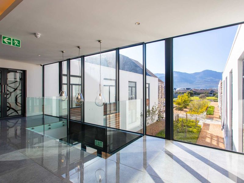 Modern, light-filled hallway featuring expansive floor-to-ceiling windows, polished tiled flooring, and a sleek glass balustrade overlooking the exterior grounds.