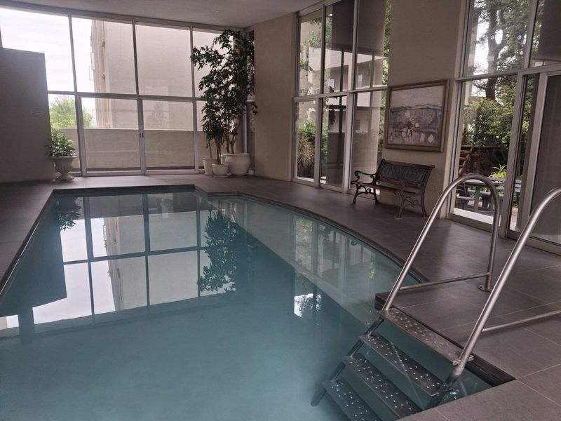 An indoor swimming pool area featuring large floor-to-ceiling windows, tiled flooring, and a stainless steel ladder, providing a bright and modern recreational space.