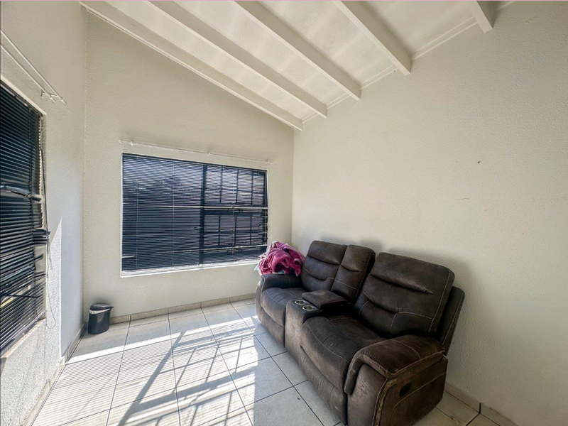 A living area featuring high ceilings with exposed beams, tiled flooring, and large windows fitted with venetian blinds.