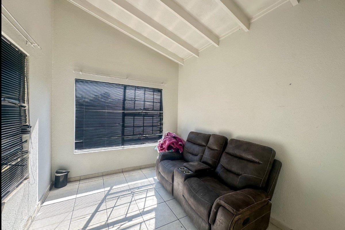 A living area featuring high ceilings with exposed beams, tiled flooring, and large windows fitted with venetian blinds.