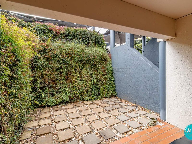 A covered patio area featuring paved flooring and a staircase, bordered by a lush, established garden wall.
