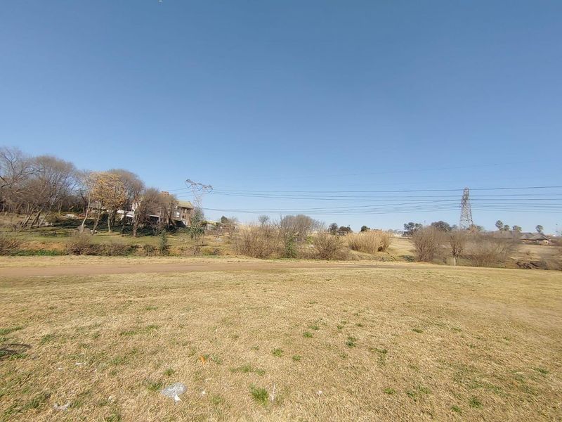 A wide-angle view of an open, grassy vacant residential plot with surrounding trees and overhead power lines visible in the distance.