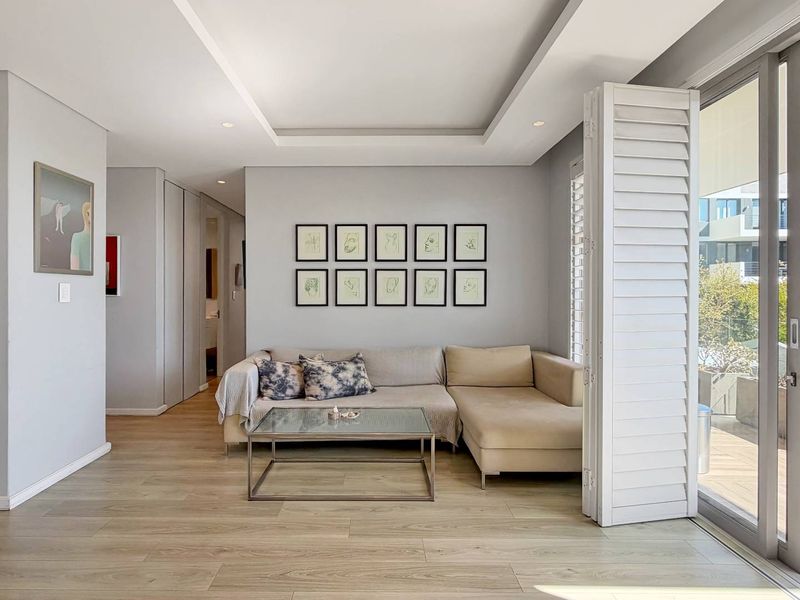 Modern living room featuring recessed ceiling lighting, light wood-look laminate flooring, and large sliding glass doors with plantation shutters opening to a balcony.