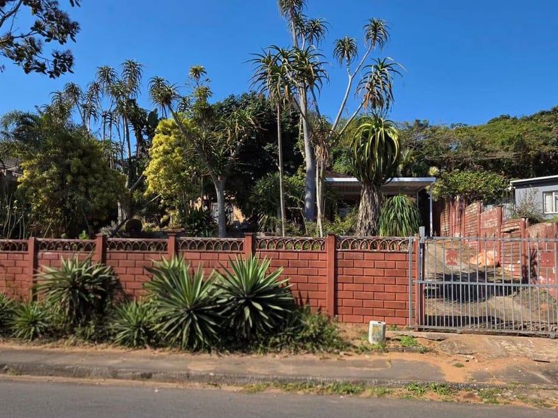 Front view of the property featuring a brick boundary wall, metal driveway gate, and established garden with mature trees.