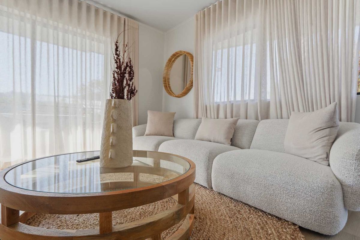 A bright and airy living room featuring large windows with floor-to-ceiling sheer curtains, providing ample natural light and a neutral, modern aesthetic.