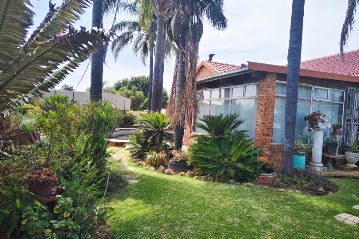A view of the rear garden featuring a brick facade, tiled roof, established palm trees, and a swimming pool area.