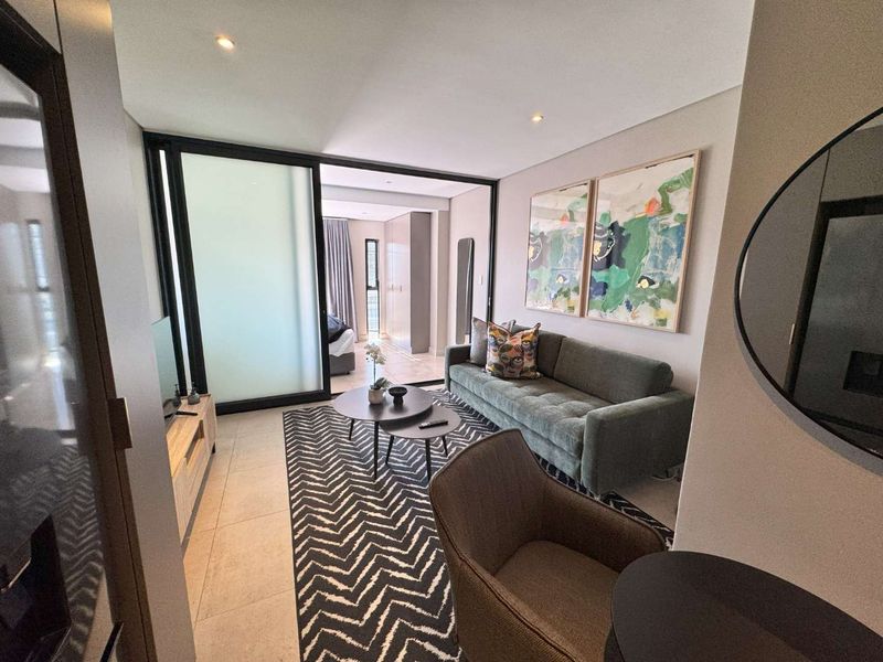 Modern living room featuring neutral tiled flooring, recessed downlighting, and a large sliding glass door providing access to the adjacent bedroom area.