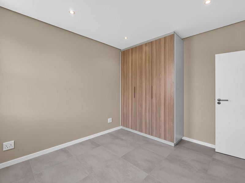 A clean, modern bedroom featuring neutral wall tones, large-format grey floor tiles, and contemporary built-in wooden cupboards with recessed ceiling lighting.