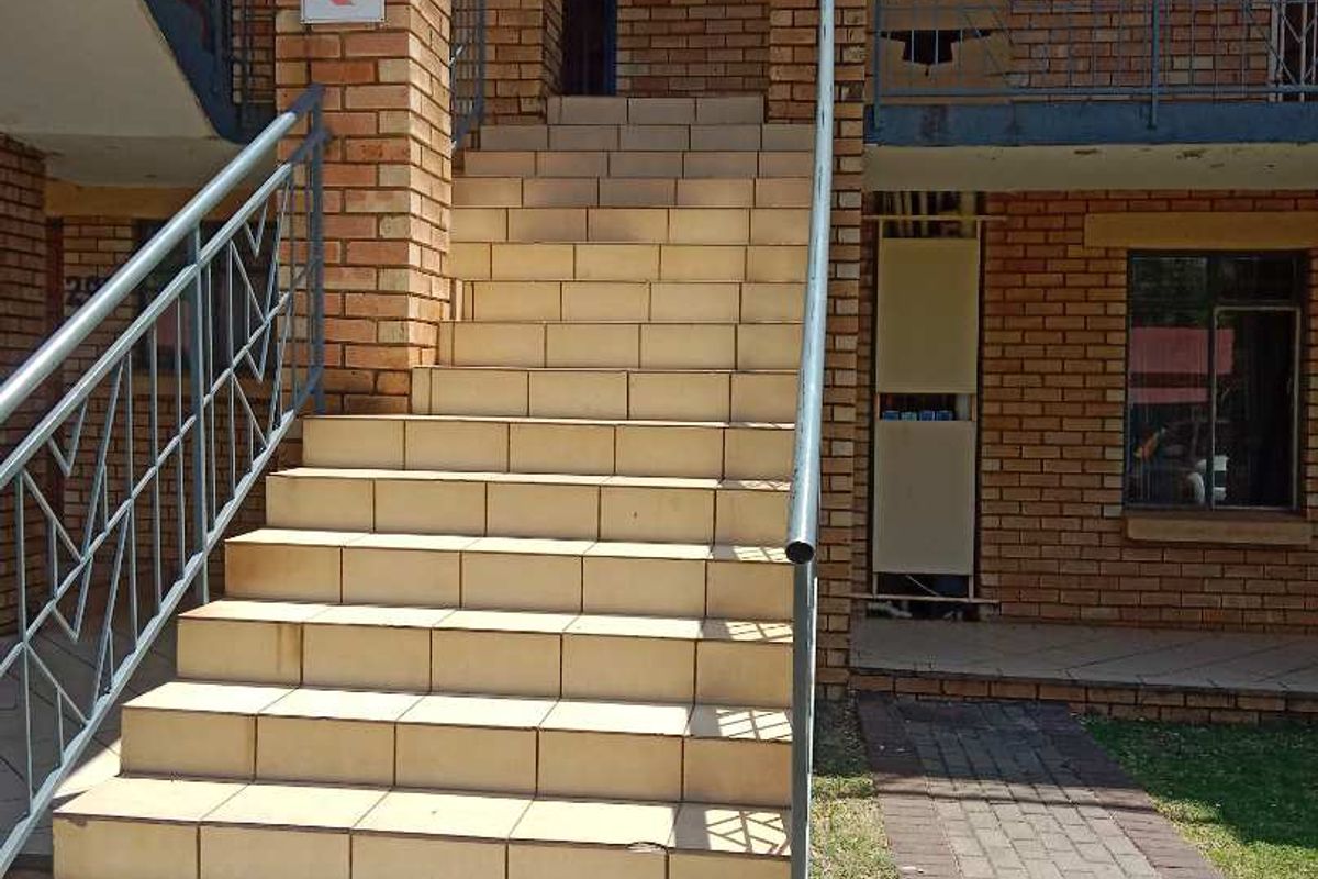 Exterior view of an apartment building entrance featuring a brick facade, tiled staircase with metal handrails, and a paved walkway.
