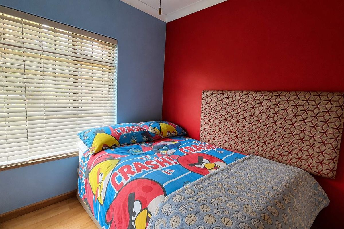A bedroom featuring laminate flooring, a window with fitted venetian blinds, and a bold accent wall.