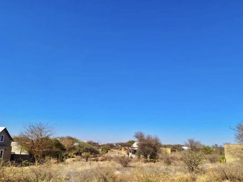 A view of a vacant residential plot featuring native vegetation and surrounding neighborhood structures under a clear blue sky.