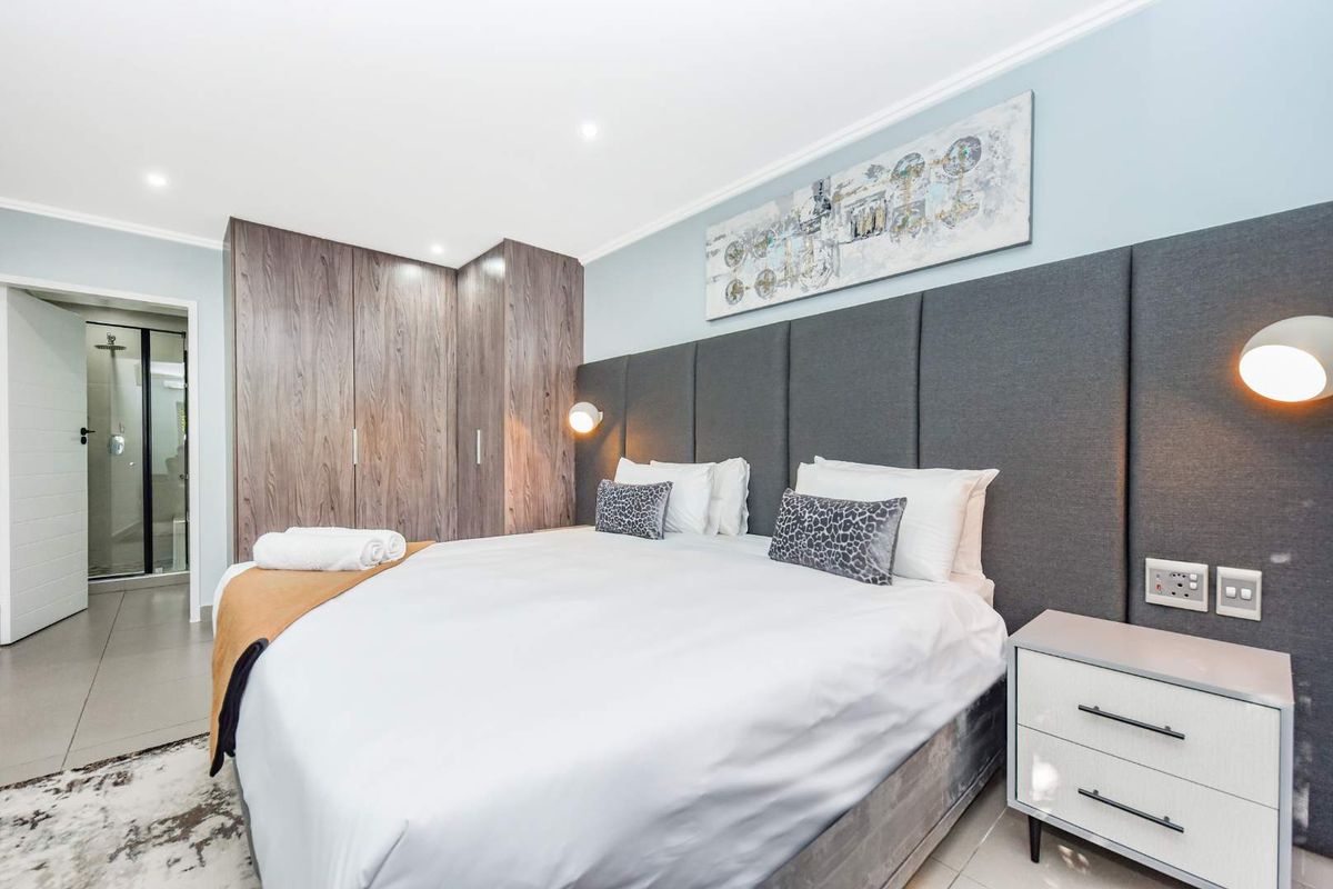 A well-maintained bedroom featuring built-in wooden cupboards, a modern upholstered headboard, recessed lighting, and direct access to an en-suite bathroom.