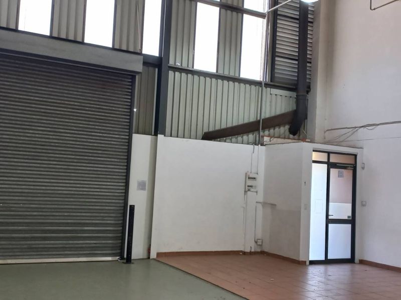 Industrial warehouse space featuring a high-clearance roller shutter door, mezzanine access staircase, and functional utility area with high ceilings.