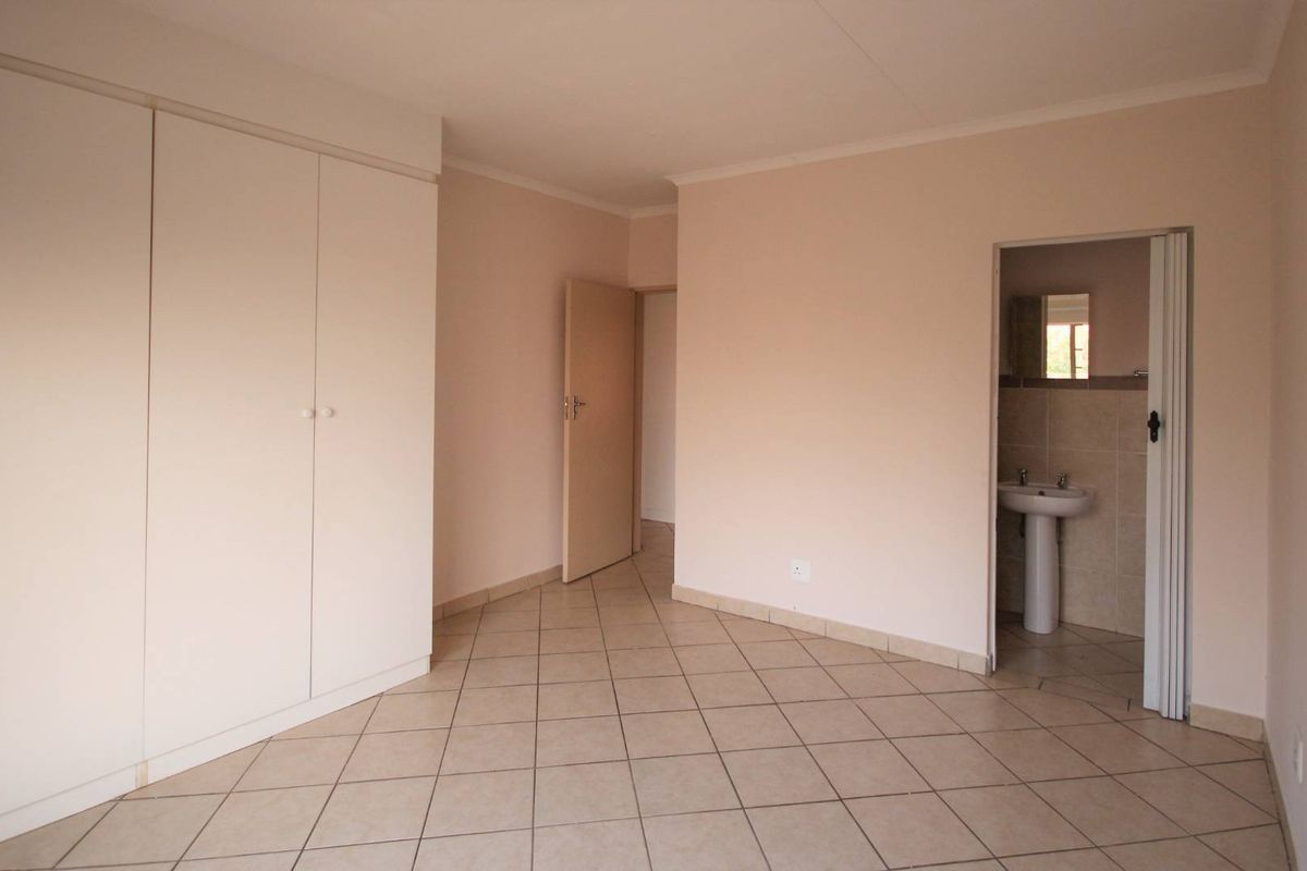 A spacious, empty bedroom featuring neutral tiled flooring, built-in cupboards, and direct access to an en-suite bathroom with a pedestal basin.