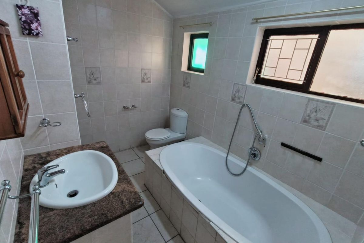 A functional bathroom featuring a built-in bathtub, a vanity with a stone countertop, and a toilet, finished with neutral wall and floor tiles.