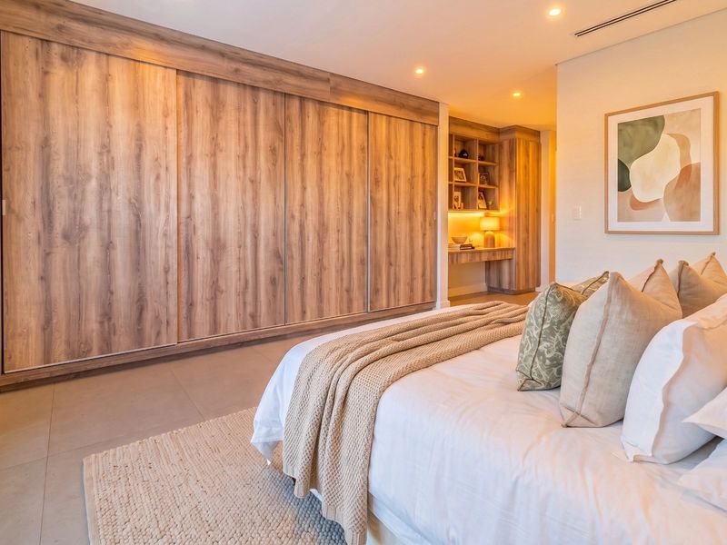 A modern bedroom featuring extensive wood-finish built-in cupboards, a dedicated built-in desk area with integrated shelving, recessed lighting, and neutral tiled flooring.