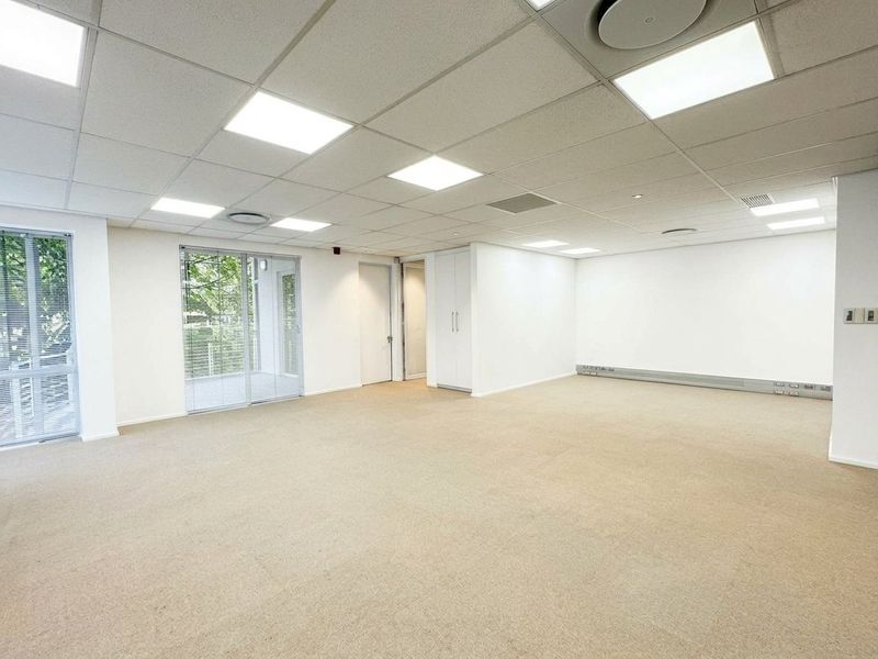 A bright, open-plan office space featuring a suspended ceiling with recessed lighting, carpeted floors, built-in storage, and access to a balcony through sliding glass doors.