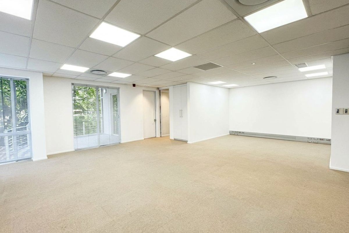 A bright, open-plan office space featuring a suspended ceiling with recessed lighting, carpeted floors, built-in storage, and access to a balcony through sliding glass doors.
