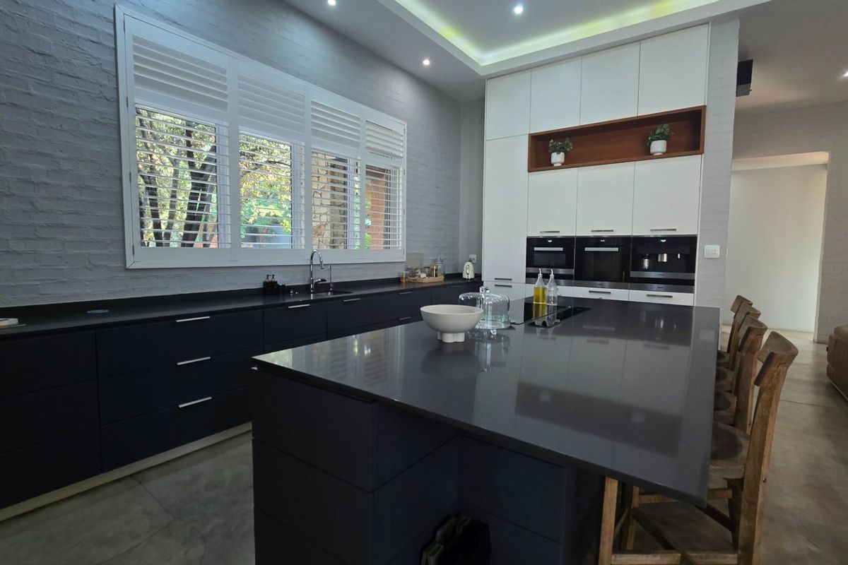 Modern kitchen featuring a large central island, dark cabinetry, stone countertops, built-in appliances, and stylish exposed brick walls with plantation shutters.