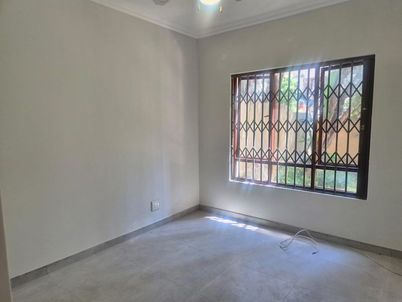 A bright, empty bedroom featuring neutral tiled flooring, coving, and a window fitted with a security gate.