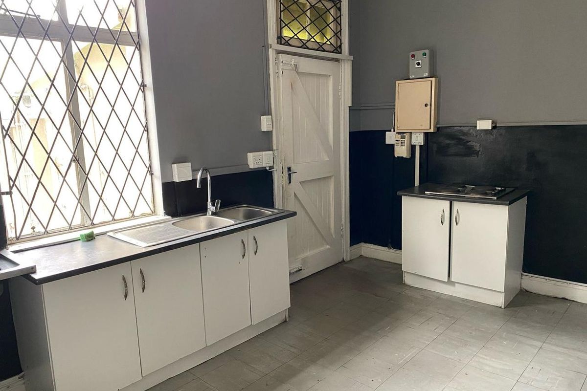 A functional kitchenette featuring white cabinetry, a stainless steel sink, and a built-in hob, set against a dark-painted wall with security-barred windows.