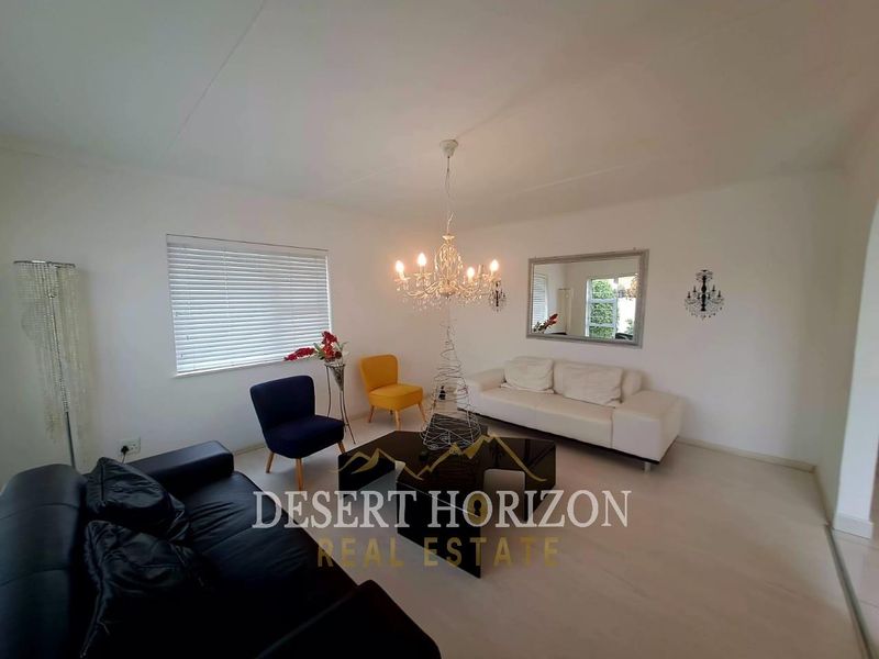 A bright living room featuring neutral flooring, venetian blinds, and a central chandelier, offering a clean and functional space for relaxation.