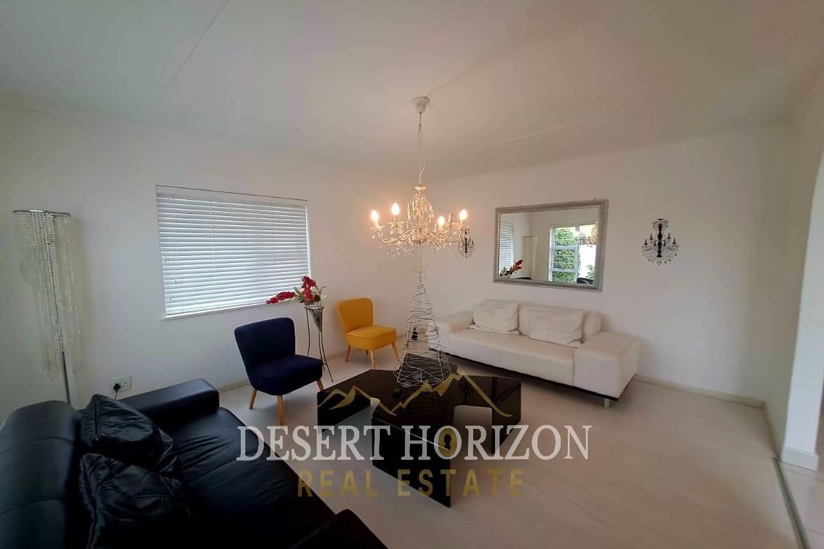 A bright living room featuring neutral flooring, venetian blinds, and a central chandelier, offering a clean and functional space for relaxation.